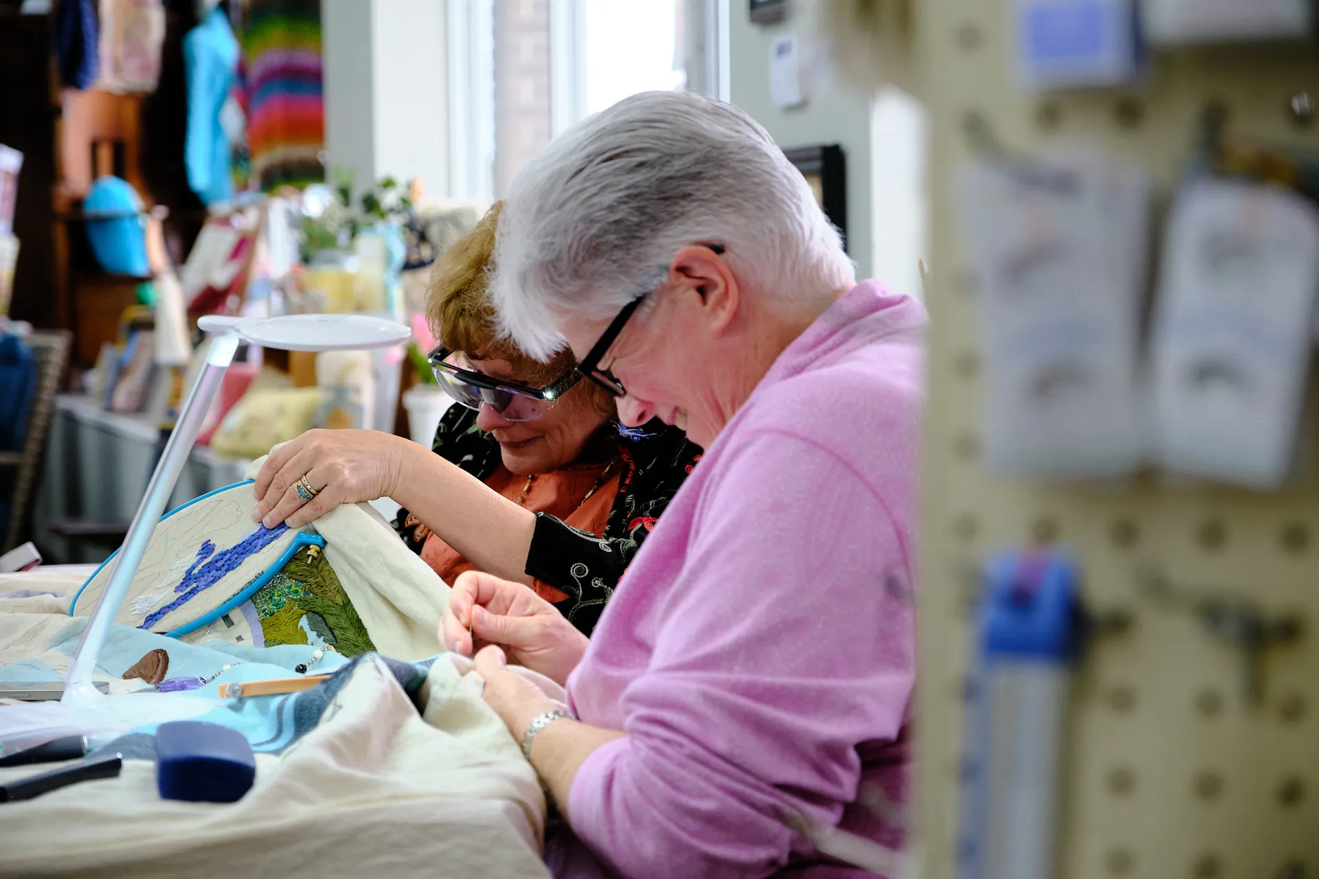 Two stitchers share a task under a work lamp, embroidering a landscape panel
