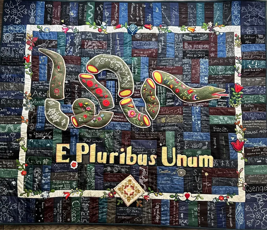 The finished quilt: the embroidered rattlesnake and the words "E Pluribus Unum" on a patchwork of signed fabric strips, inside a cream border of stitched flowers